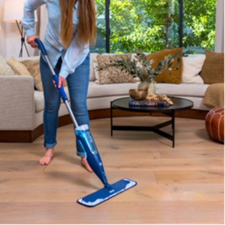 Mujer limpia el suelo de madera con un mop con cabezal azul en una sala iluminada.